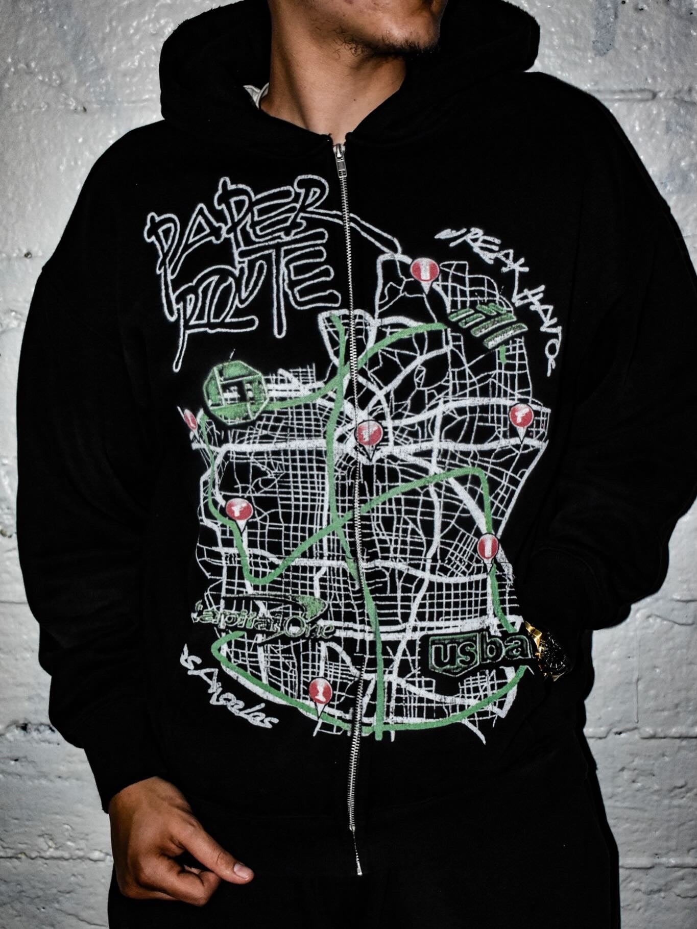 Person wearing a black hoodie with a map design and text on a gray brick wall background